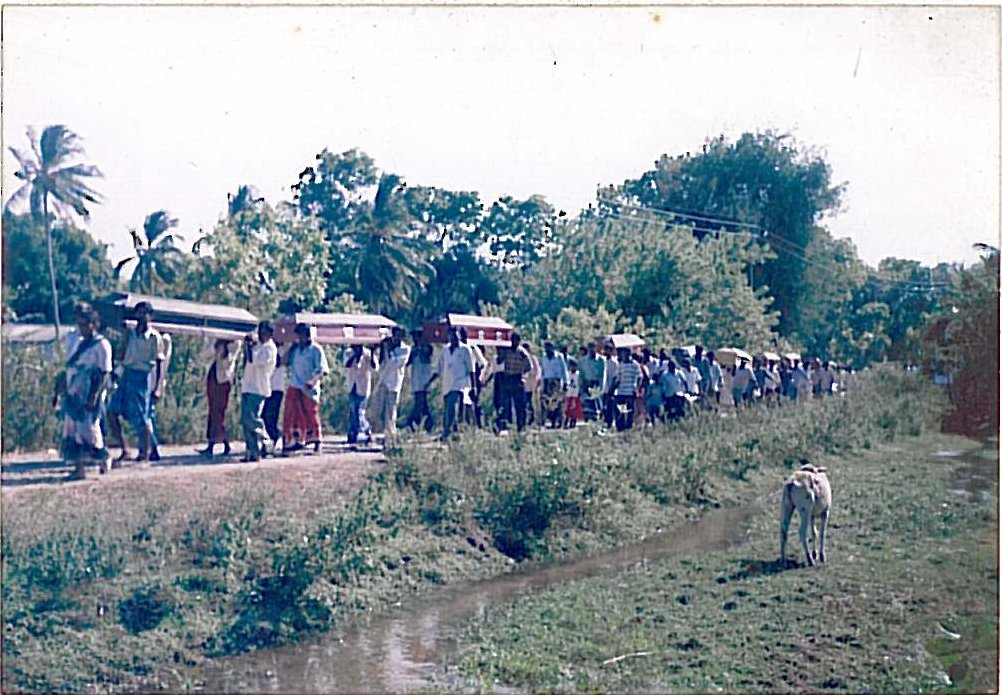 Mourners on a rural road carry a line of coffins past a canal, followed by a long procession of villagers