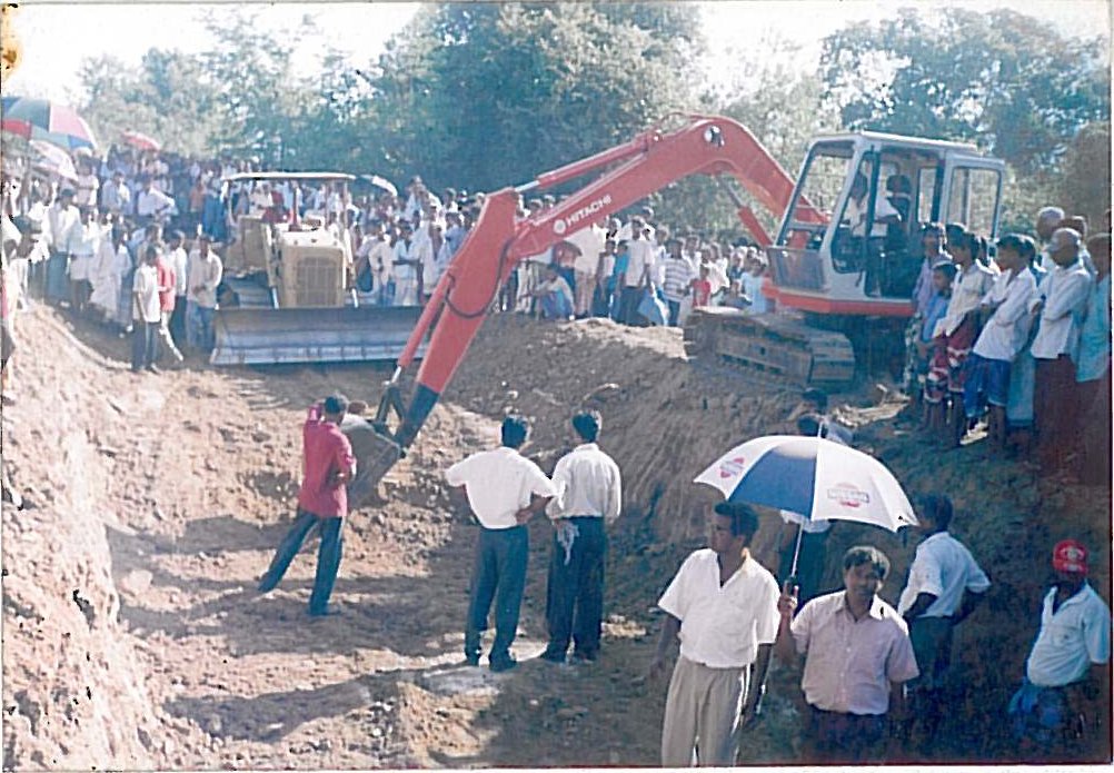 An excavator digs a long trench while crowds of people, some holding umbrellas, watch from the edges